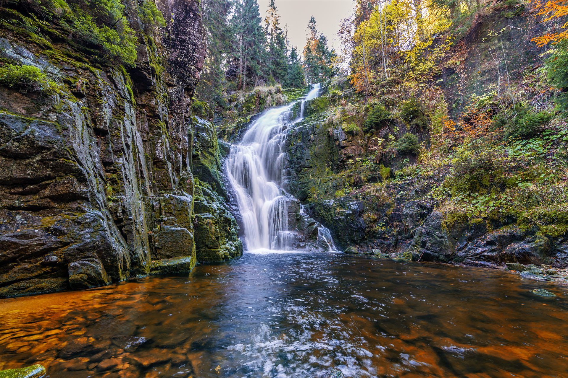 Wodospad spadający po mszystych skałach do ciemno-brązowego nurtu, otoczony jesiennymi drzewami