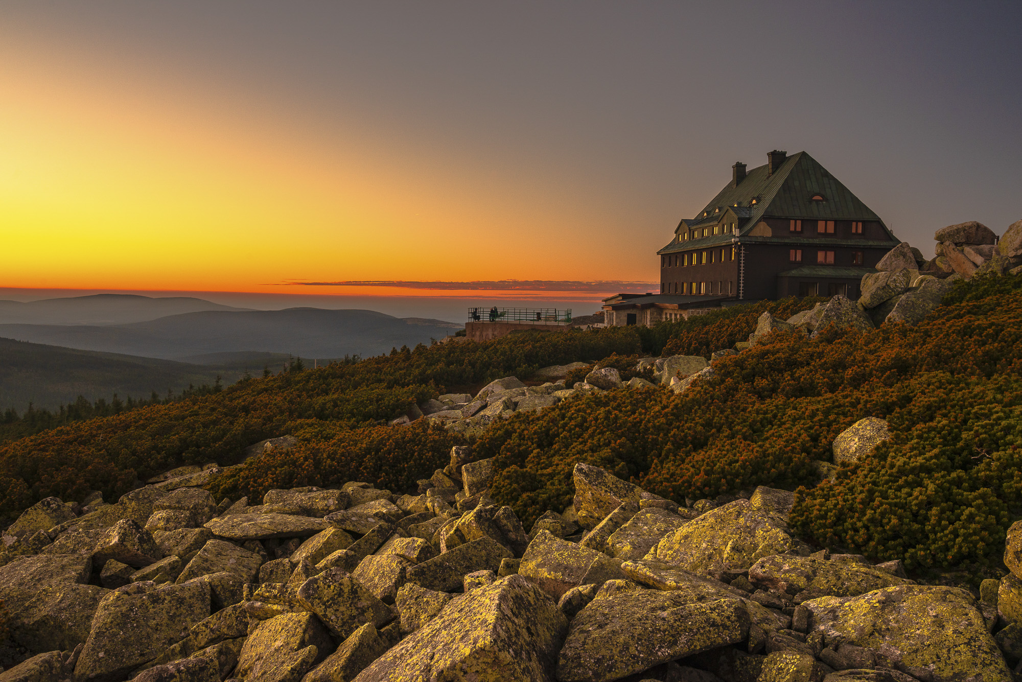 Hotel na skalistym stoku, porośniętym krzewami, przy pomarańczowym zachodzie nad falistymi górami