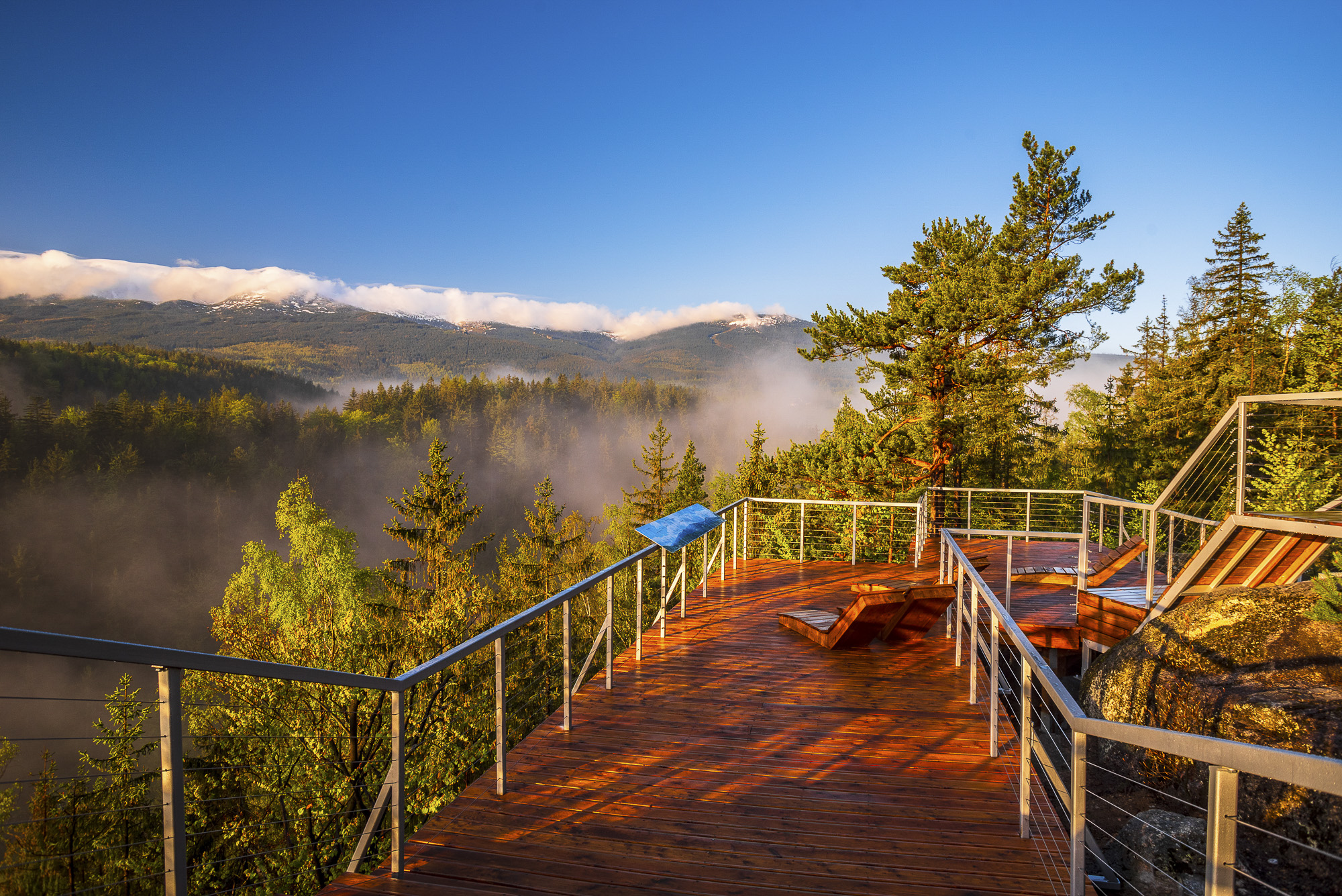 Drewniany taras widokowy z leżakami nad mglistym lasem i ośnieżonymi górami pod błękitnym niebem