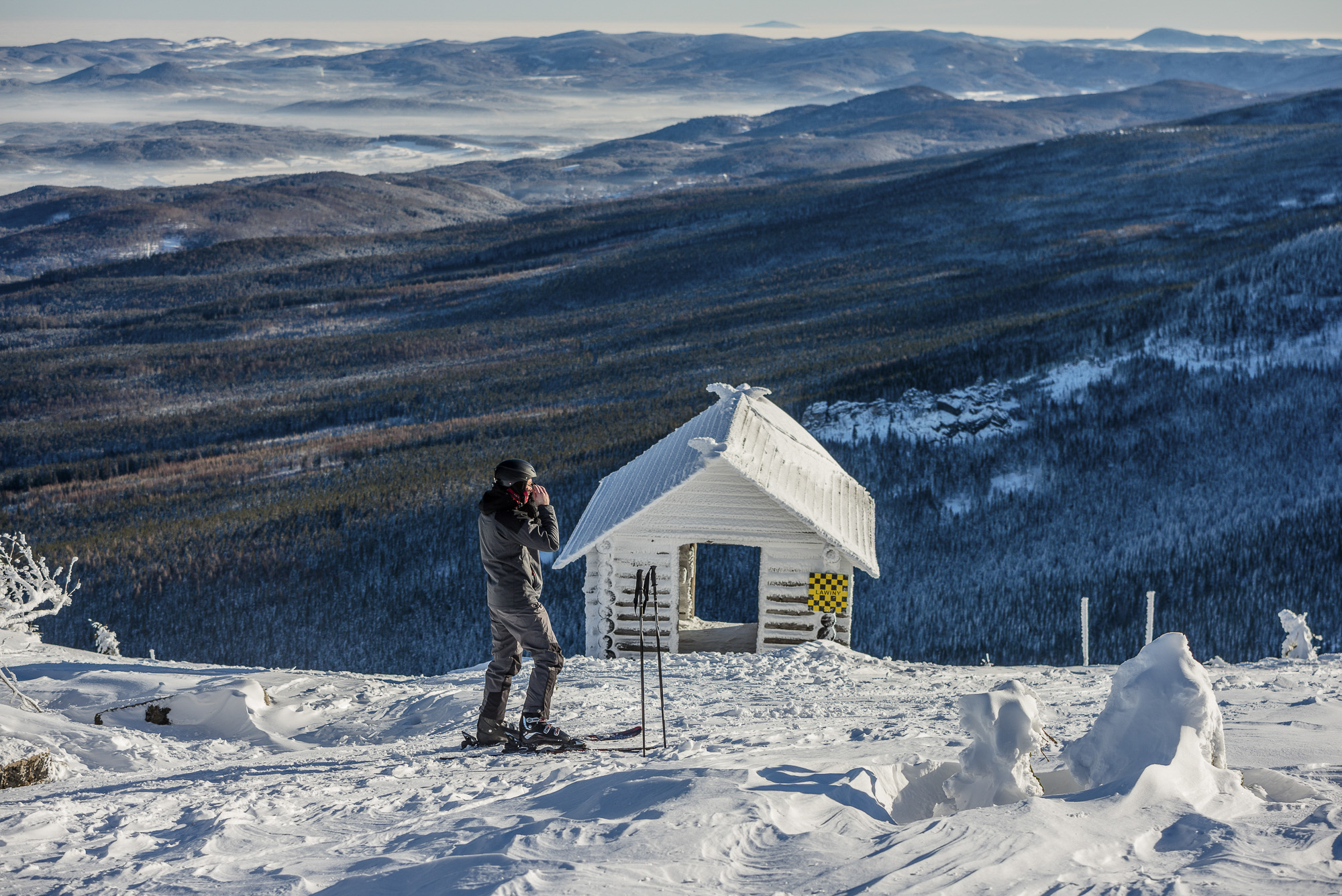 Narciarz przy nartach obok zaśnieżonej chatki na szczycie, w tle rozległe niebiesko-zielone góry