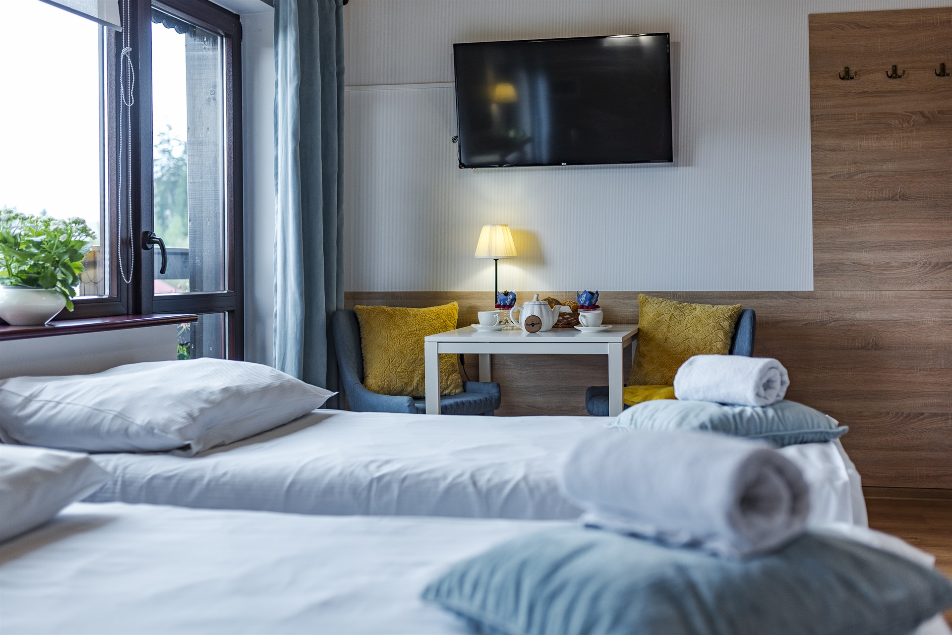 Dwie łóżka z białą pościelą i zwiniętymi ręcznikami, stolik z lampą, telewizor i żółte poduszki przy oknie