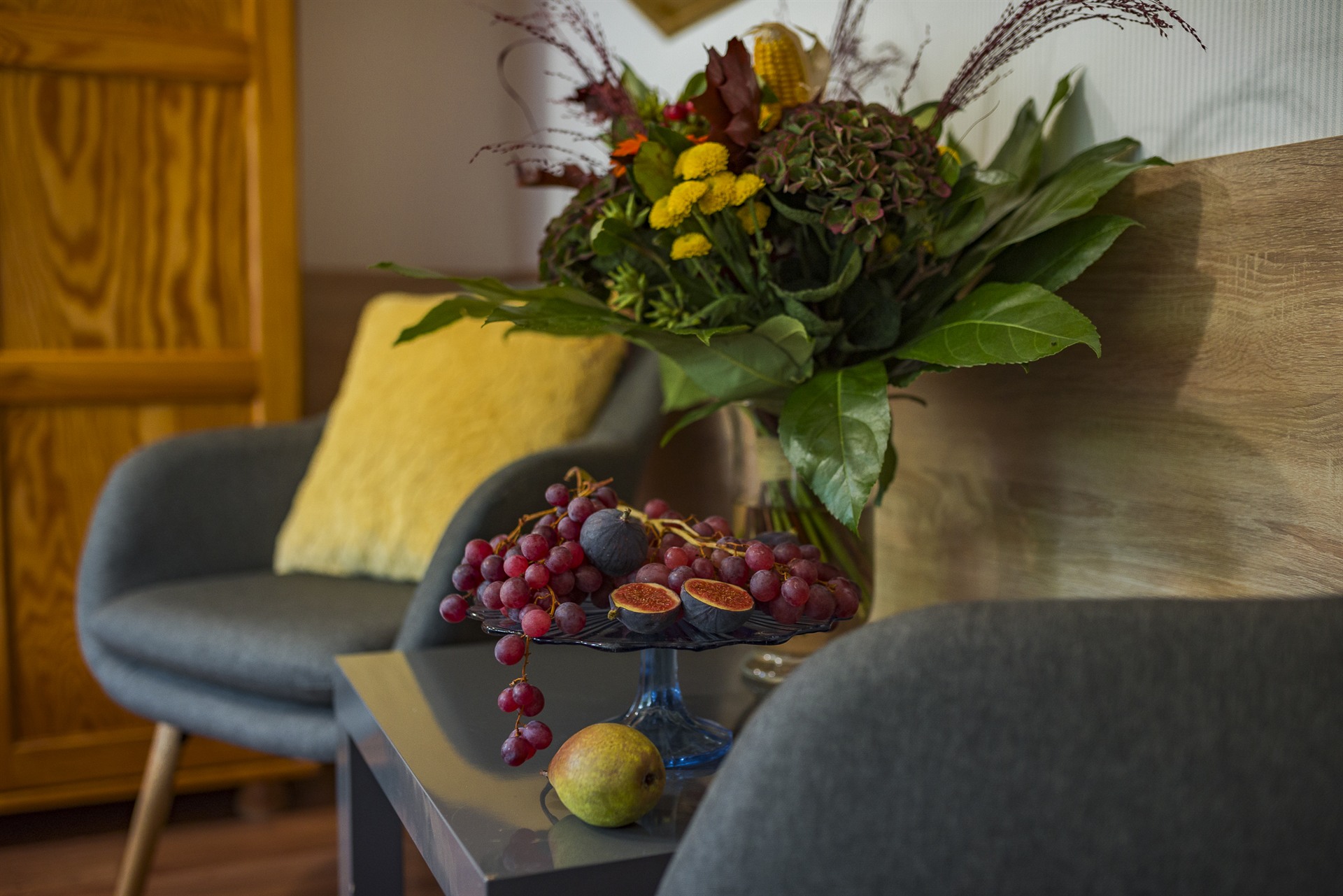 Kiść czerwonych winogron i przekrojone figi na niebieskiej paterze obok gruszki, w tle duży bukiet i szare fotele