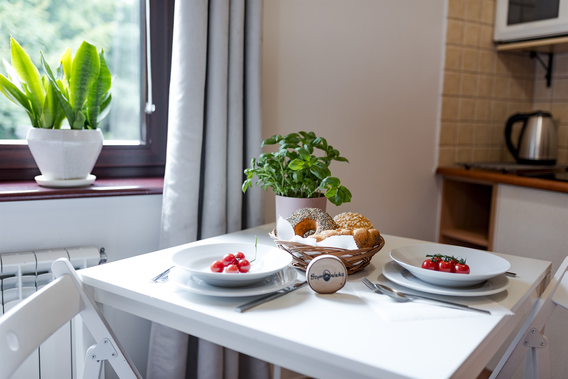 Biały stół nakryty dla dwóch osób z talerzami z pomidorkami, koszykiem pieczywa i doniczką bazylii