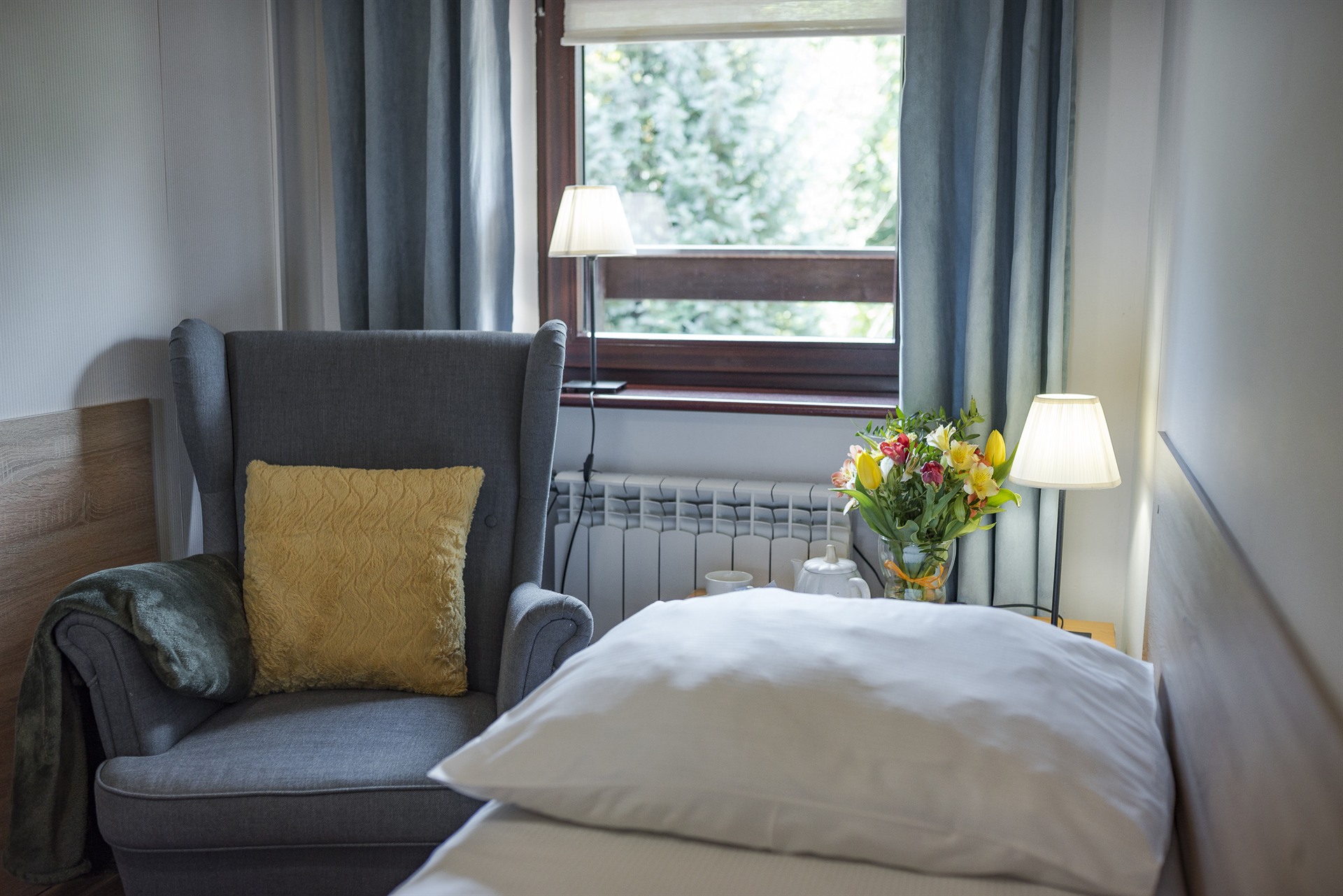 Szary fotel z żółtą poduszką przy łóżku, okno z zasłonami, stolik z lampą i bukietem kolorowych kwiatów