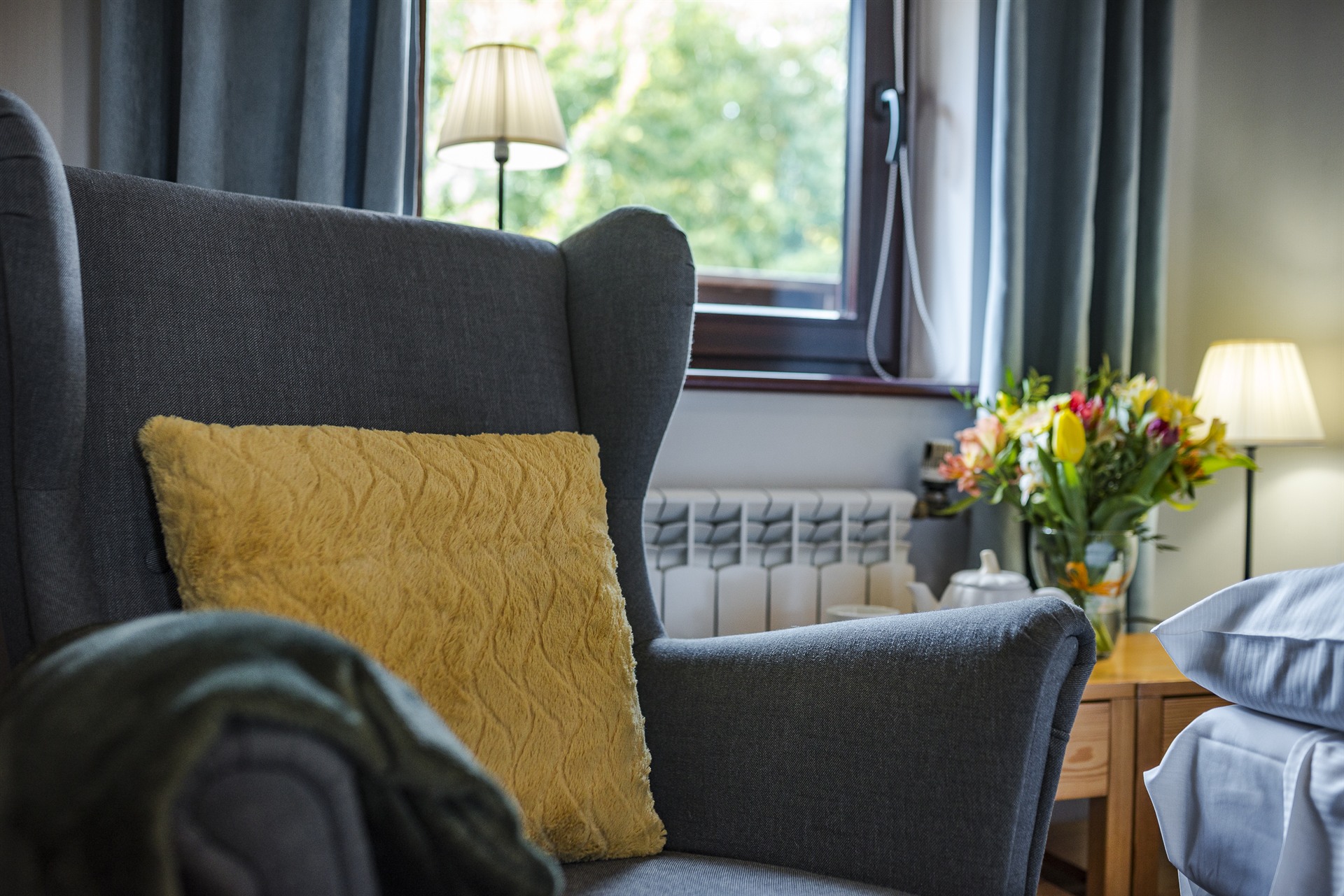 Szary fotel z żółtą poduszką przy oknie, lampy i bukiet kwiatów na stoliku