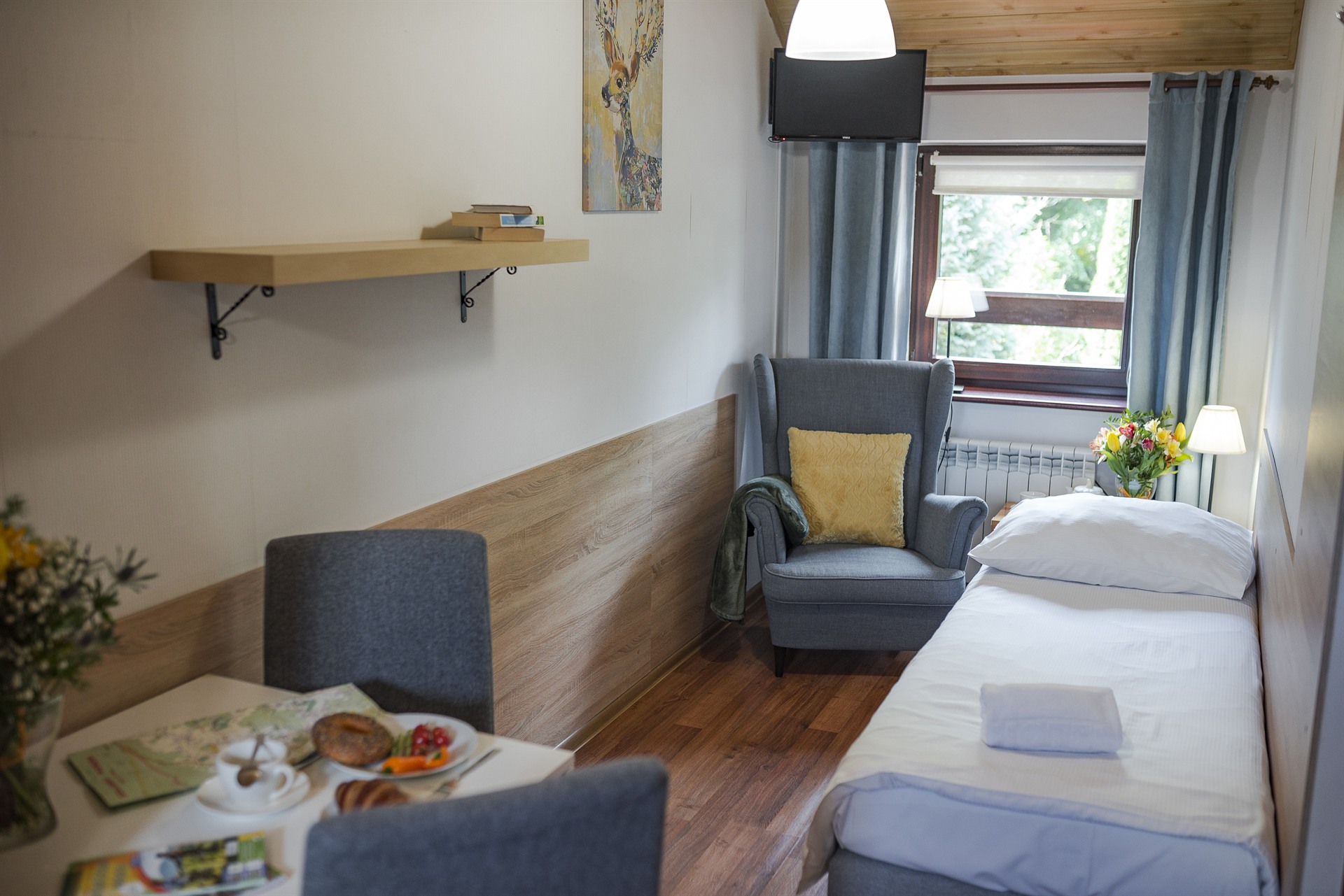 Kameralny pokój z pojedynczym łóżkiem, szarym fotelem z żółtą poduszką, stołem ze śniadaniem i bukietem
