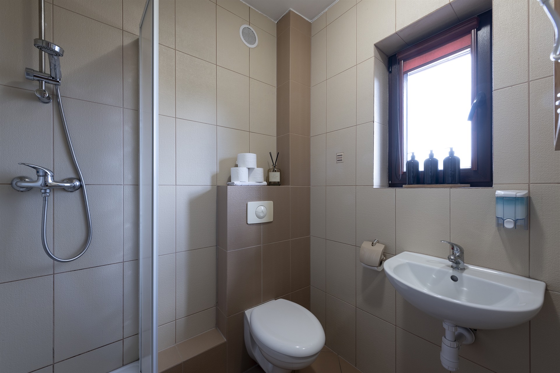 Mała łazienka z wiszącą toaletą, umywalką po prawej, kabiną prysznicową po lewej i oknem z trzema czarnymi butelkami