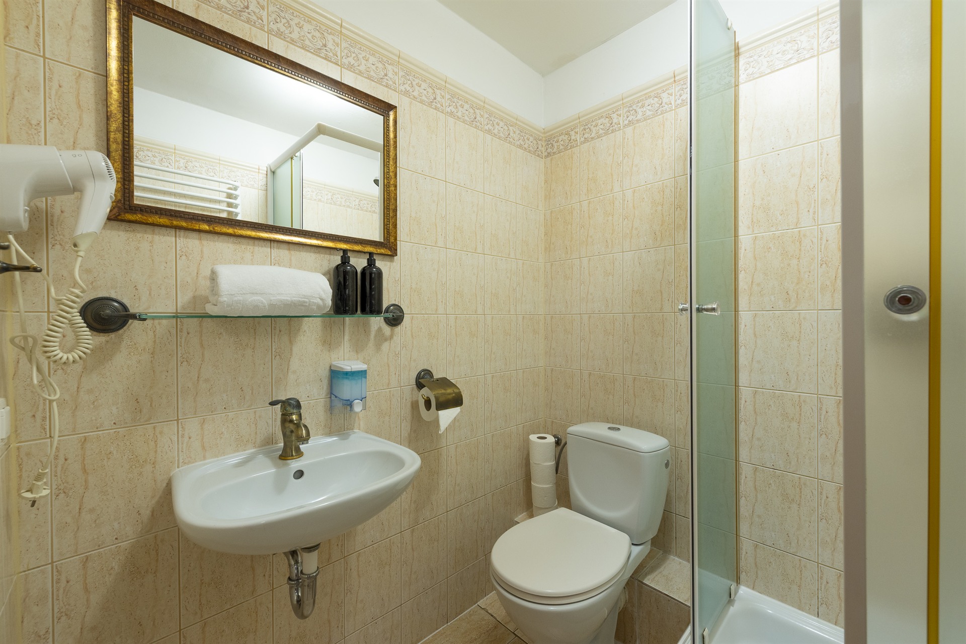 Mała łazienka z beżowymi płytkami, umywalką z mosiężną baterią, złotym lustrem, ręcznikiem, czarnymi butelkami, toaletą i prysznicem