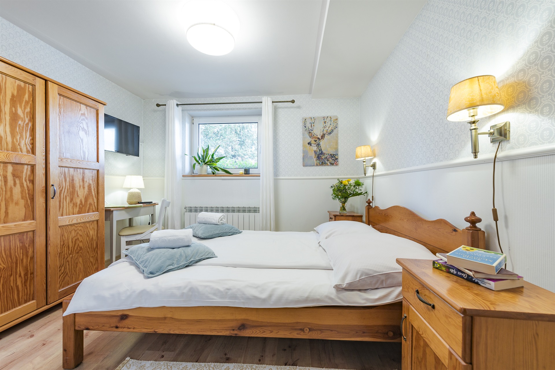 Drewniane łóżko z białą pościelą i niebieskimi poduszkami w przytulnej sypialni z szafą, lampami i rośliną przy oknie