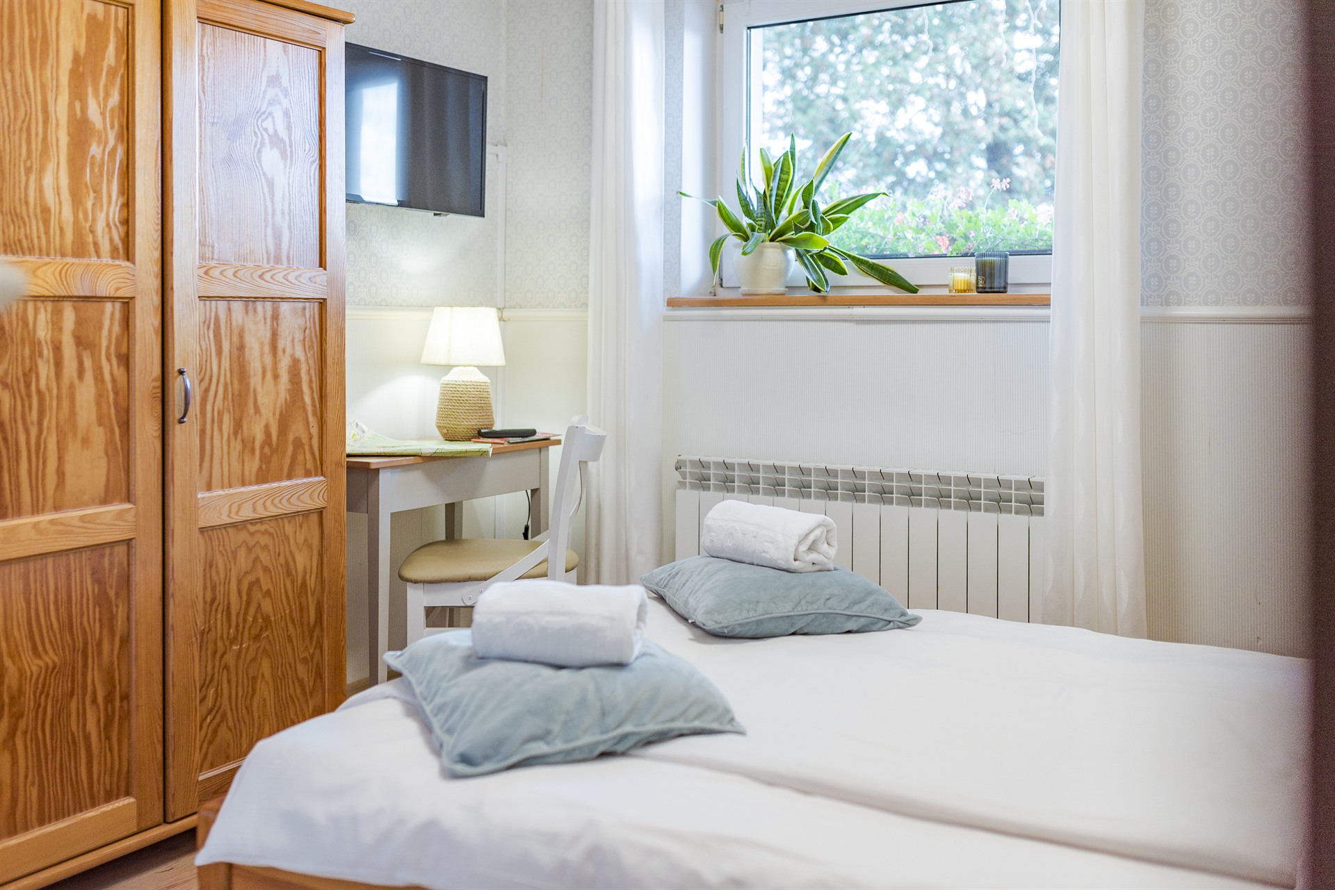 Łóżko z niebieskimi poduszkami i zwiniętymi ręcznikami, obok drewniana szafa i biurko z lampą przy oknie z rośliną