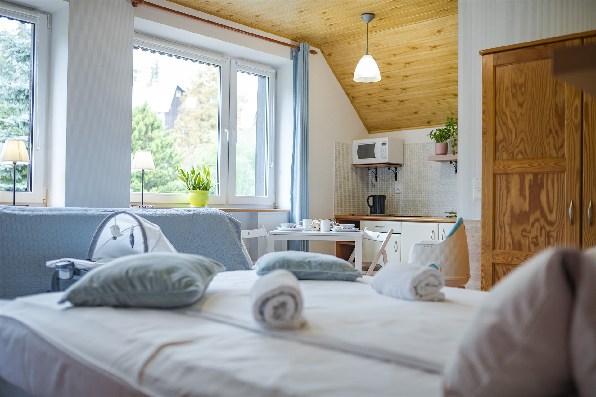 Słoneczne studio poddasze z łóżkiem i zrolowanymi ręcznikami, oknem z doniczką i małą kuchnią z mikrofalą