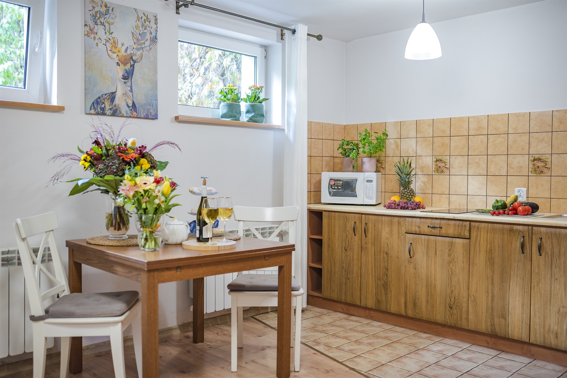 Jasna kuchnia z drewnianym stołem i bukietem kwiatów, obrazem jelenia, mikrofalówką i owocami