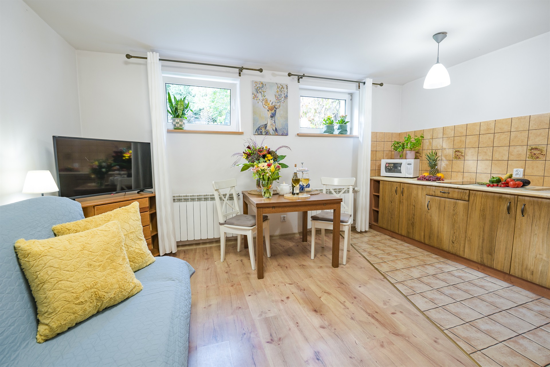 Kompaktowy salon z niebieską sofą i żółtymi poduszkami, drewniany stół z bukietem i kuchnia w tle