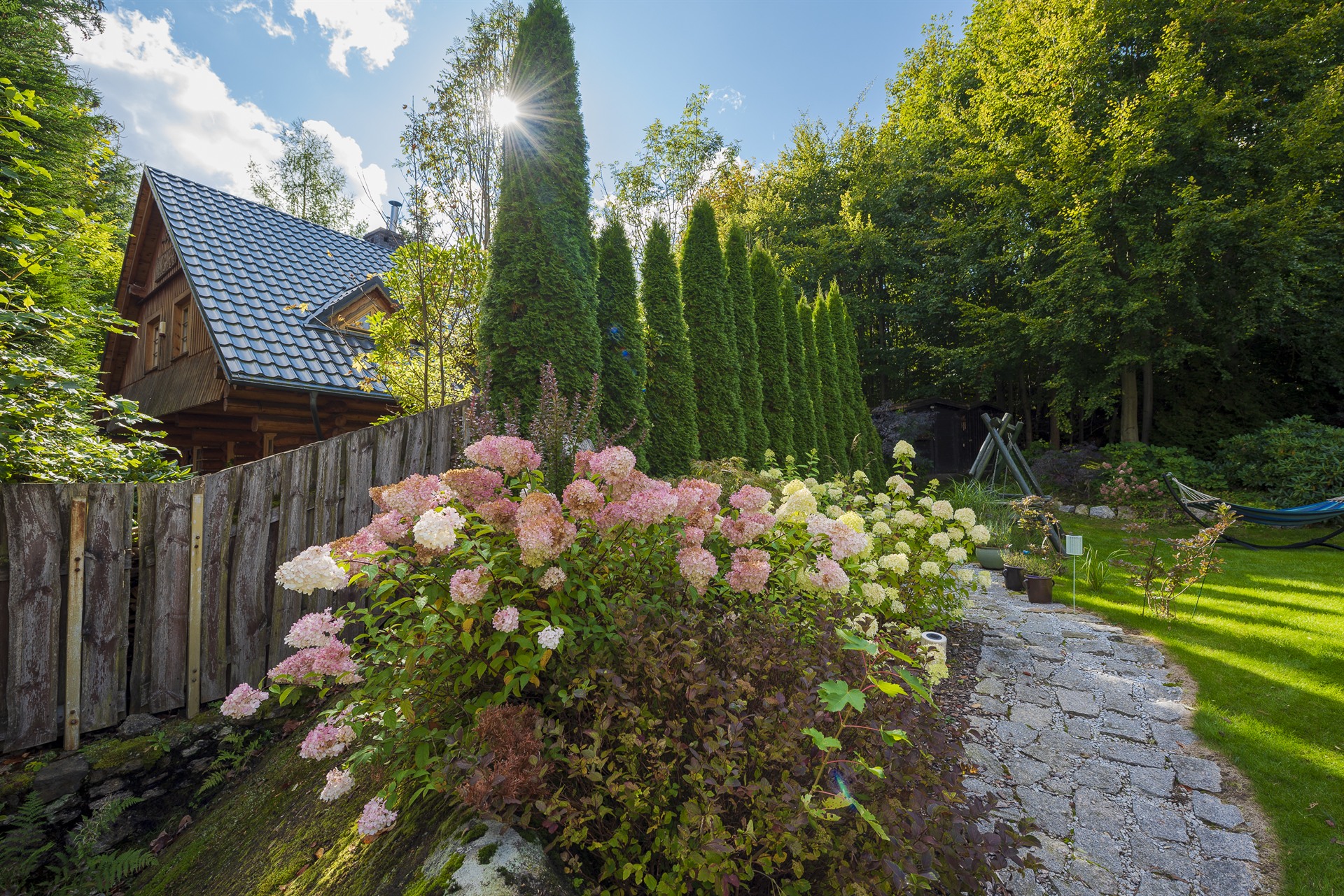 Drewniany domek z niebieskim dachem, przed nim rabata różowo-białych hortensji i kamienna ścieżka między tujami