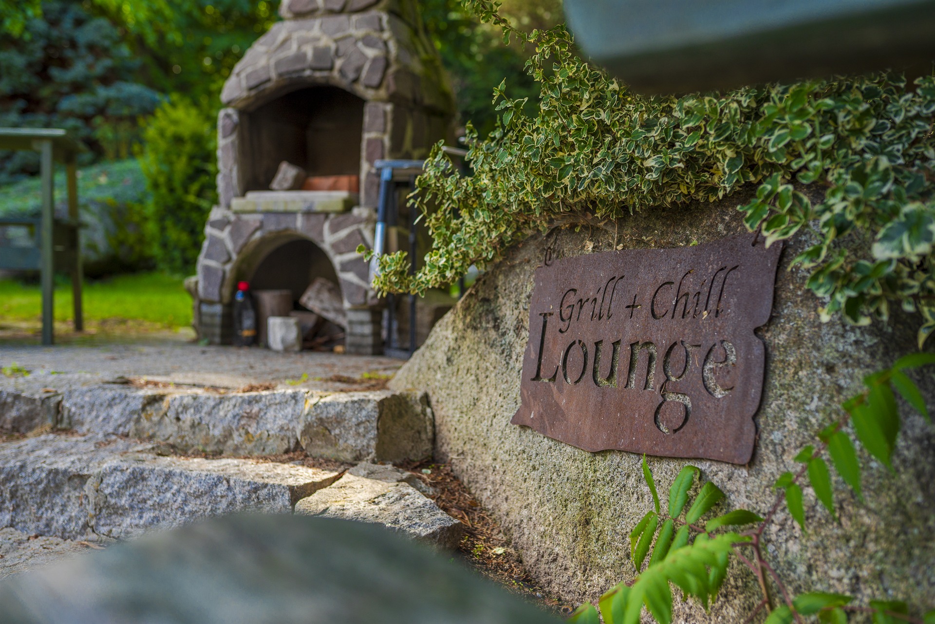 Rustykalny metalowy szyld Grill + Chill Lounge na skale obok kamiennego grilla w zielonym ogrodzie
