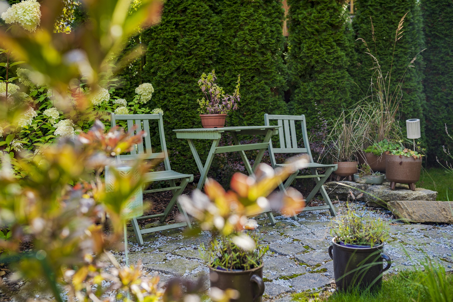 Zielony drewniany stolik z dwoma krzesłami w kamiennym patio, doniczki i białe hortensje w tle