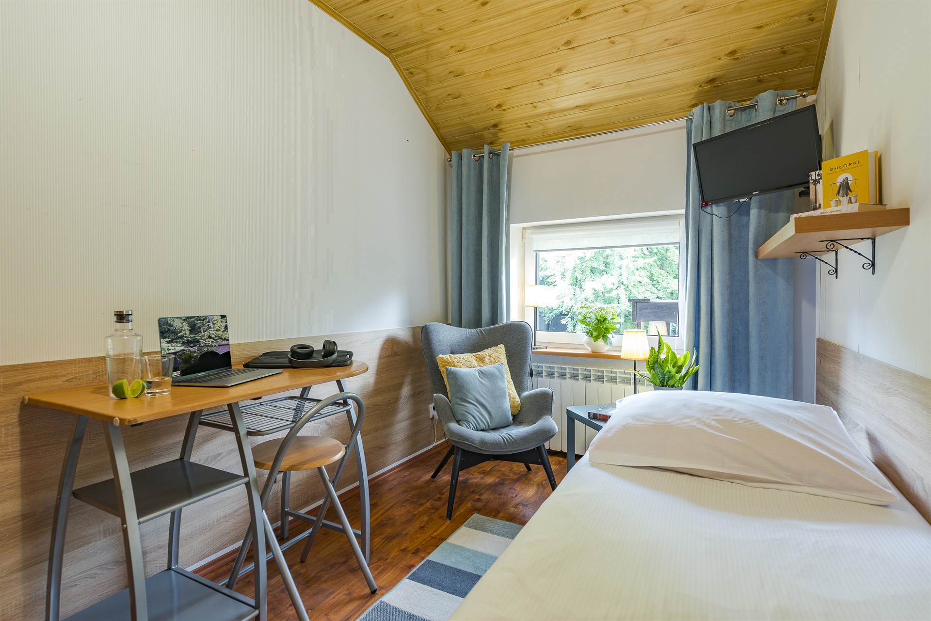 Jasny pokój z pojedynczym łóżkiem, biurkiem z laptopem, fotelem przy oknie i roślinami