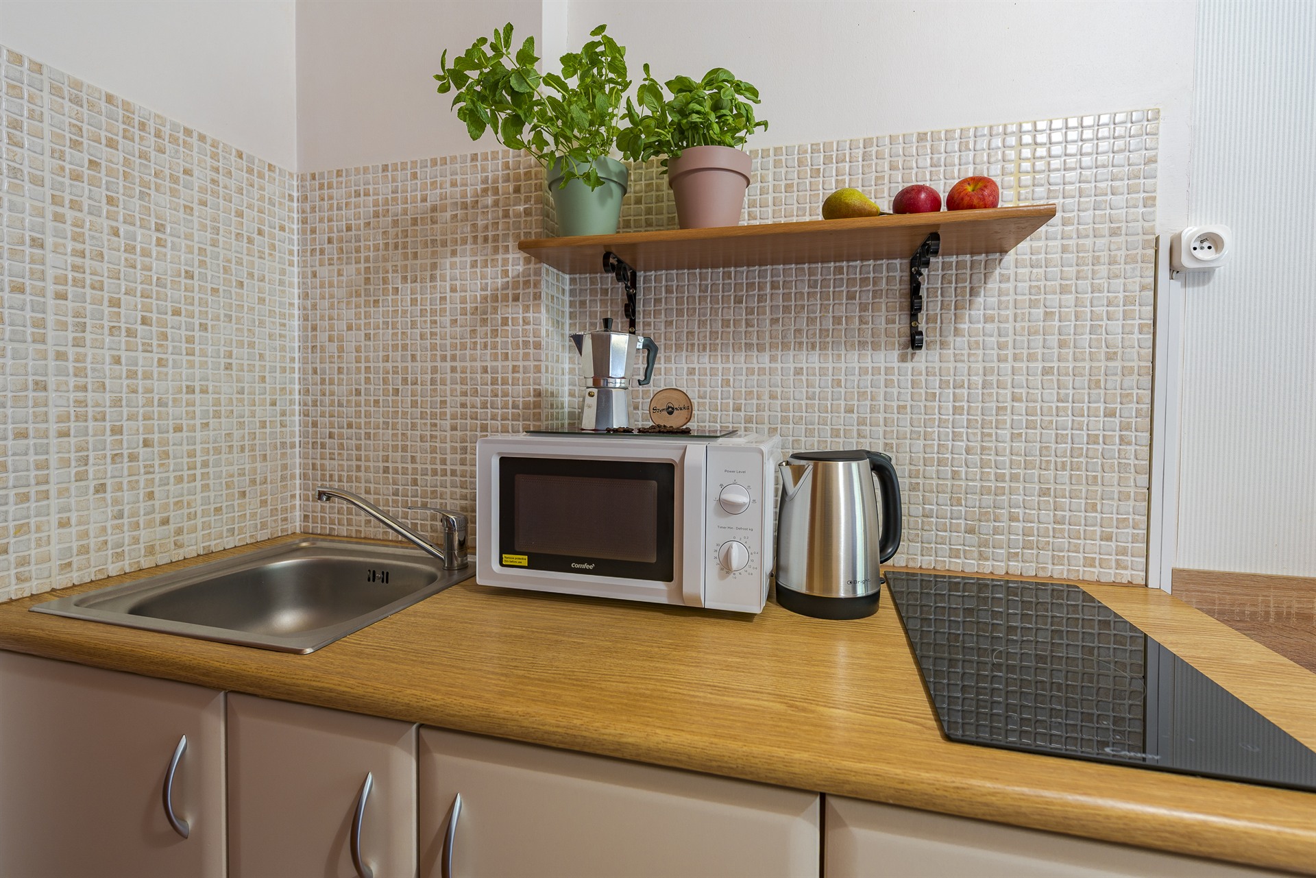 Kuchenny blat z mikrofalówką, czajnikiem, dwoma doniczkami z ziołami na półce i owocami