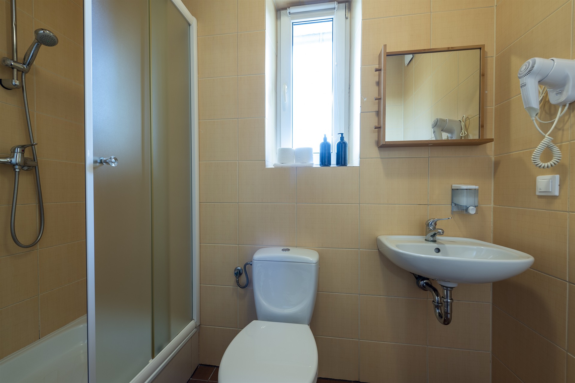Beżowa łazienka z kabiną prysznicową, toaletą, umywalką, lustrem i oknem z niebieskimi butelkami