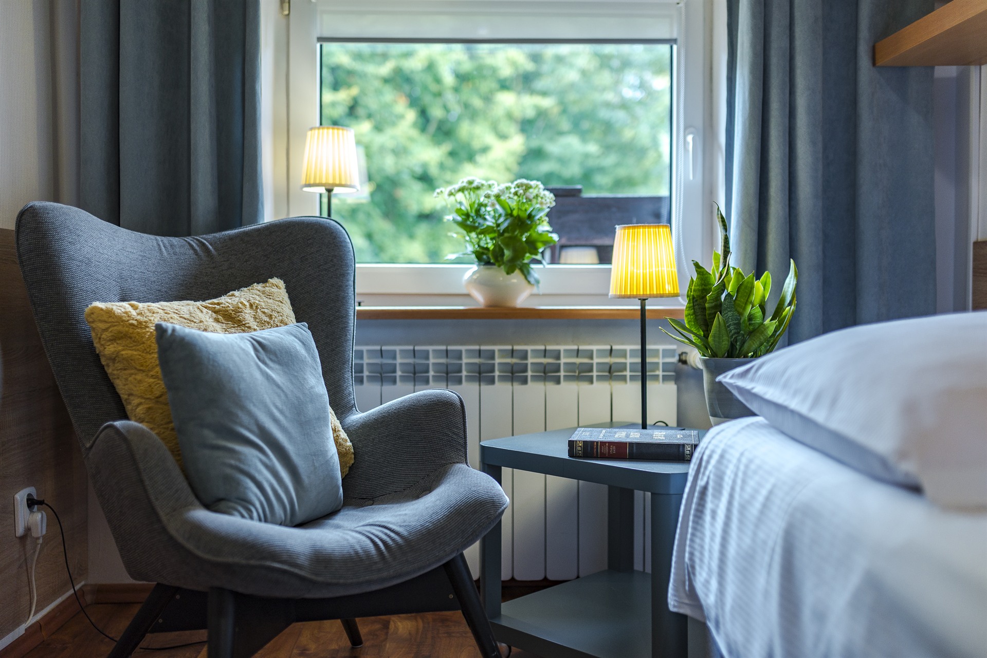 Szary fotel z poduszkami przy stoliku z lampą i roślinami przy oknie z zielonym widokiem