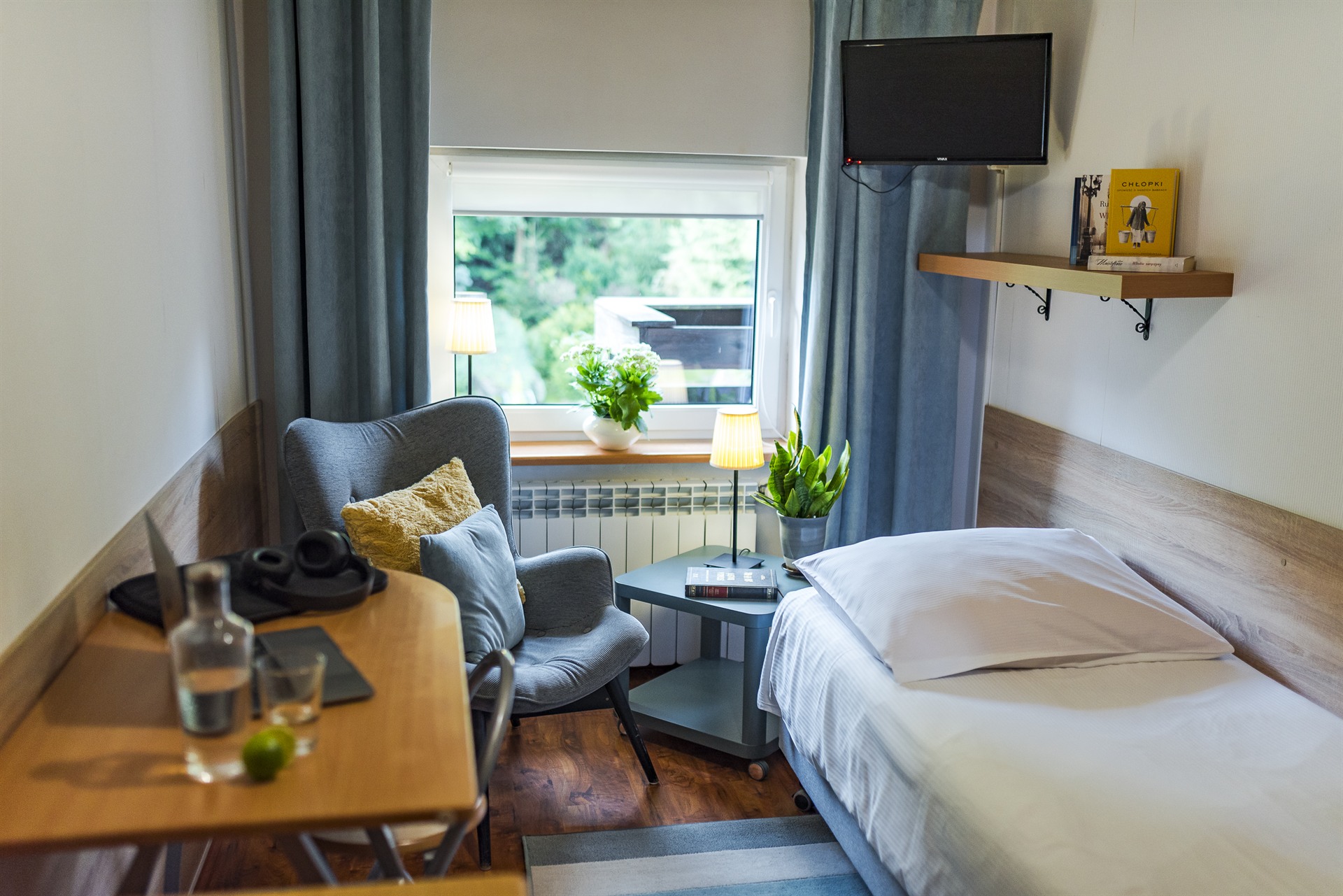 Mały pokój z pojedynczym łóżkiem, szarą fotelą z żółtą poduszką, stolikiem z lampą i roślinami przy oknie