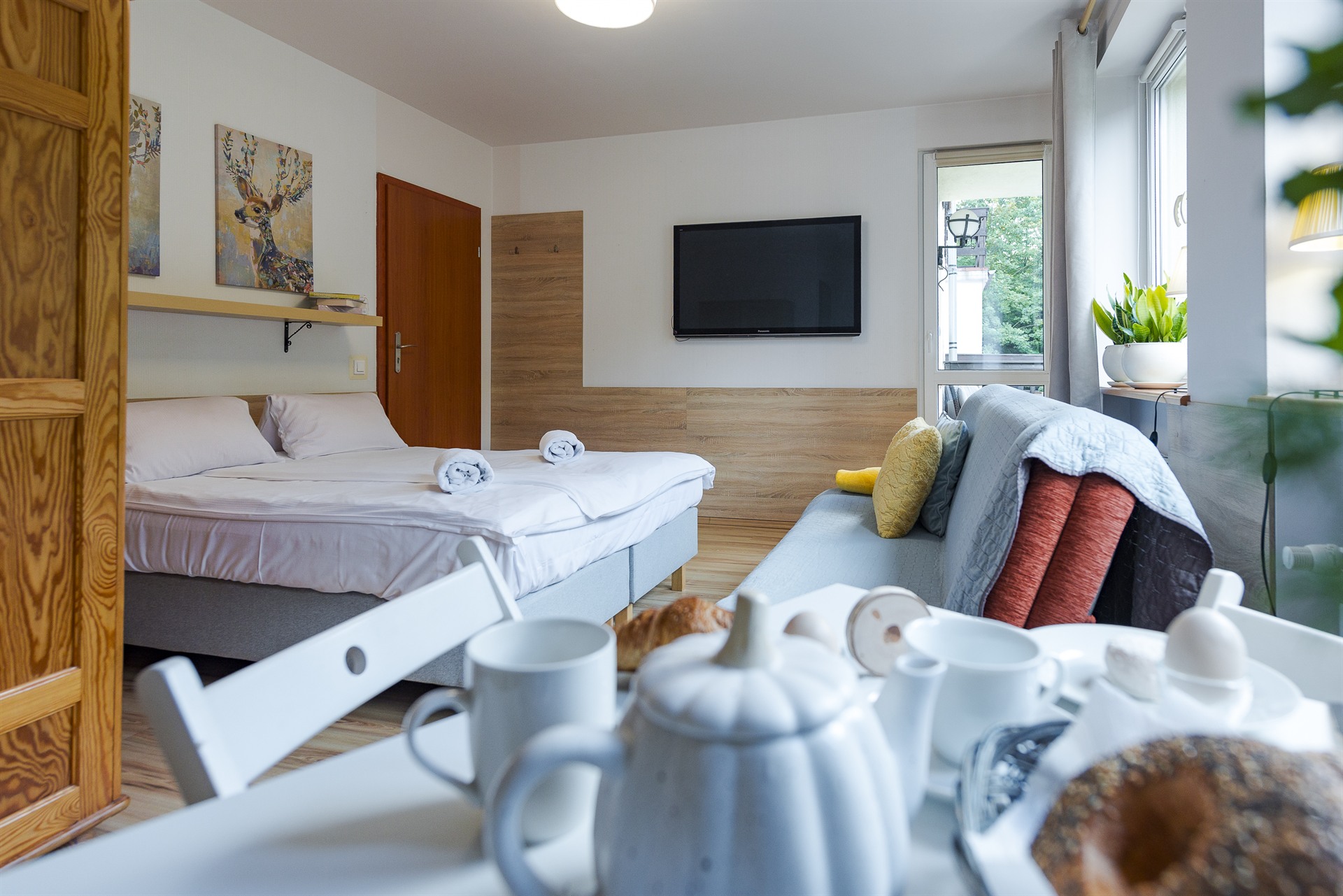 Sypialnio‑pokój z łóżkiem i zwiniętymi ręcznikami, telewizorem na ścianie i sofą z kolorowymi poduszkami