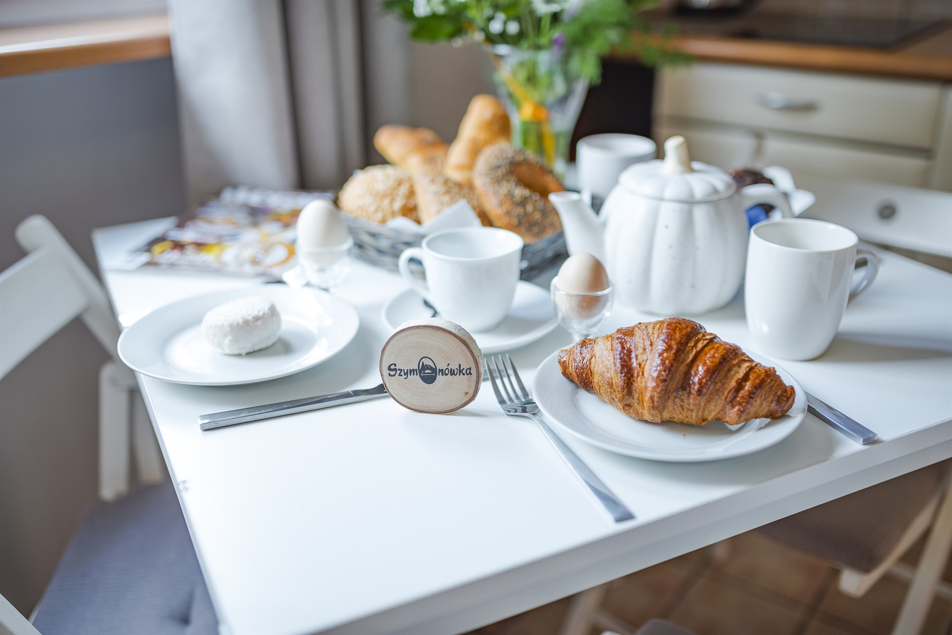 Złocisty croissant na talerzu, biała filiżanka i dzbanek obok jajek w kieliszkach i koszyka z pieczywem