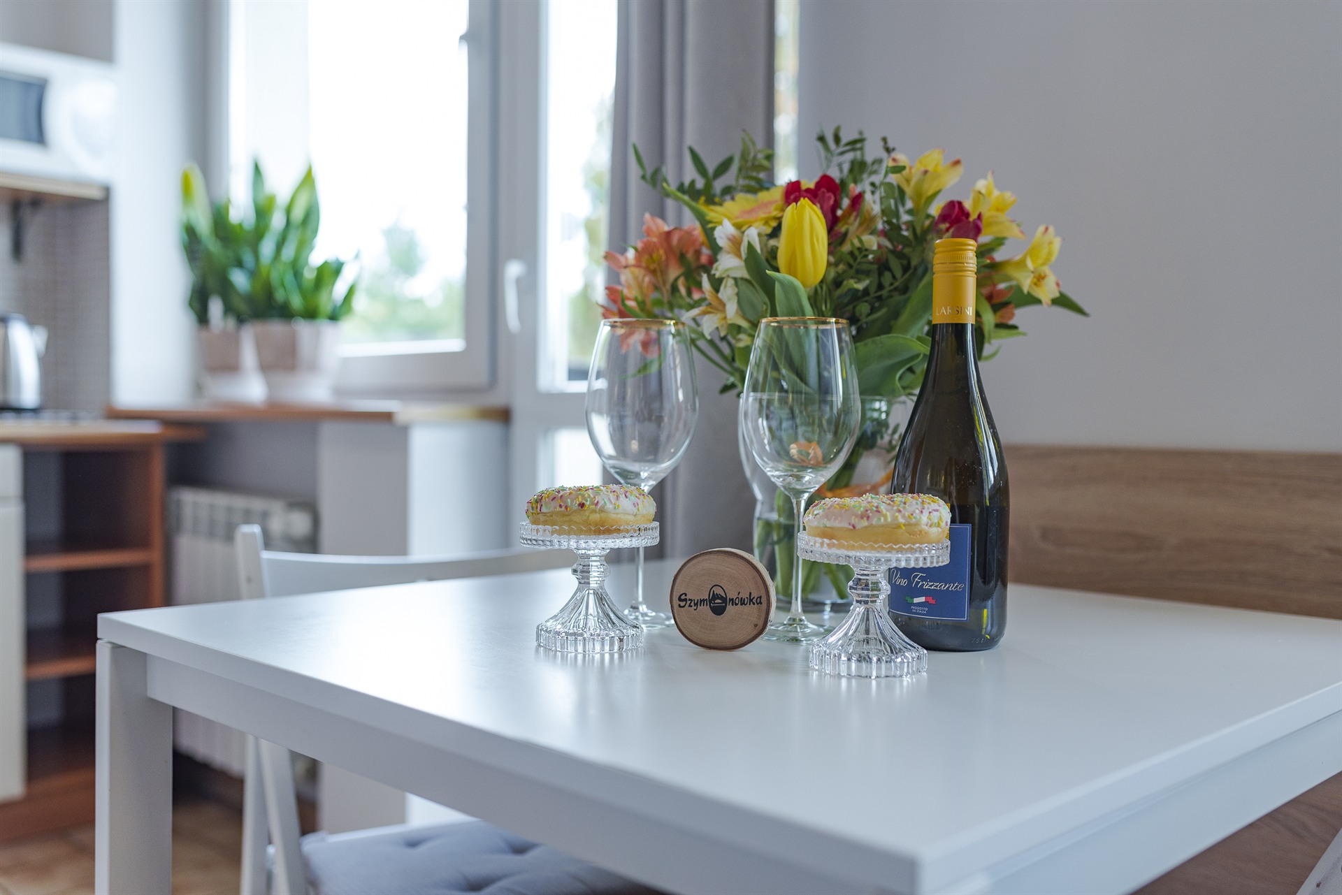 Butelka wina, dwa kieliszki, bukiet tulipanów i dwa kremowe pączki z posypką na szklanych podstawkach