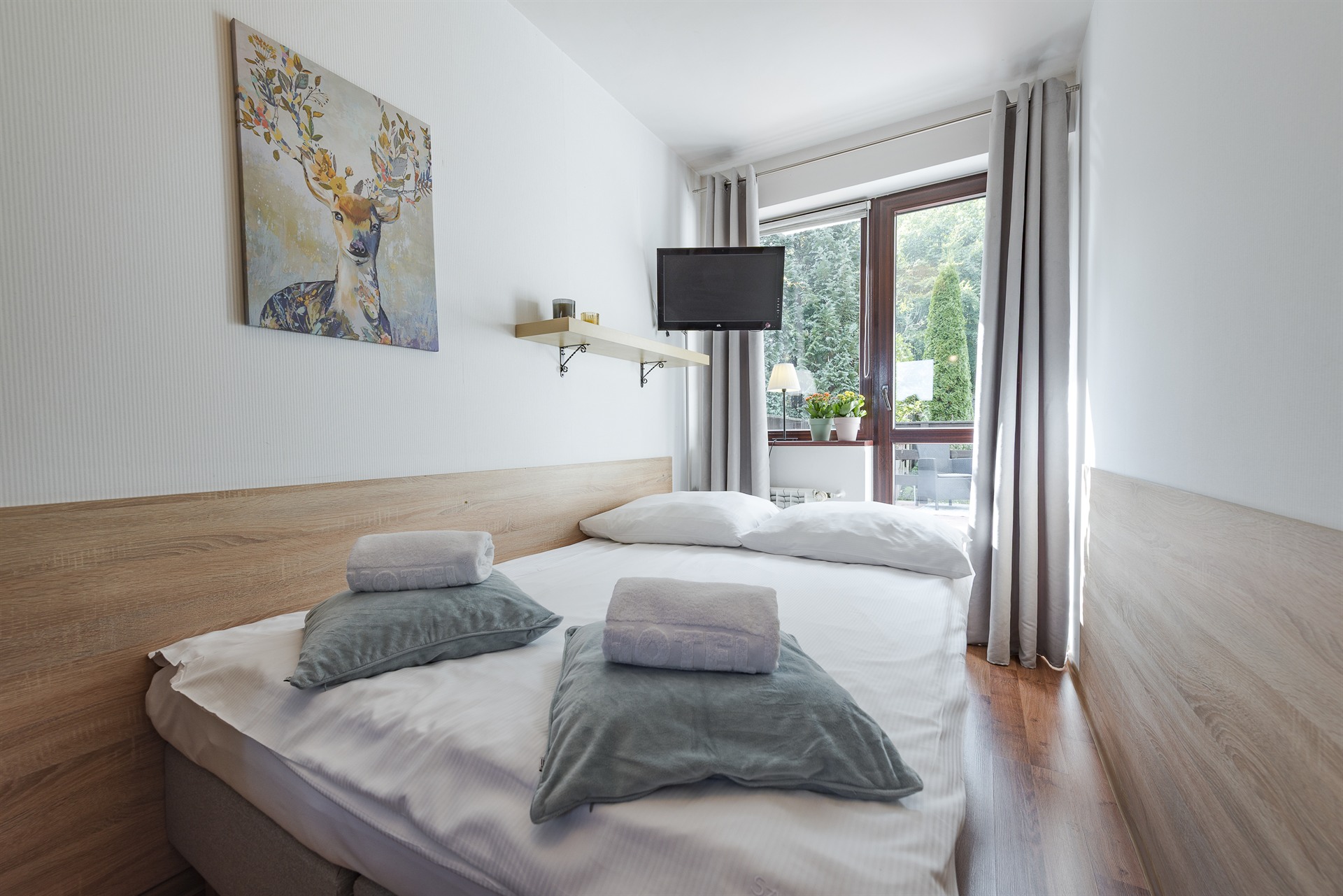 Jasna sypialnia z podwójnym łóżkiem, drewnianym zagłówkiem, poduszkami i ręcznikami, okno z zielonym widokiem