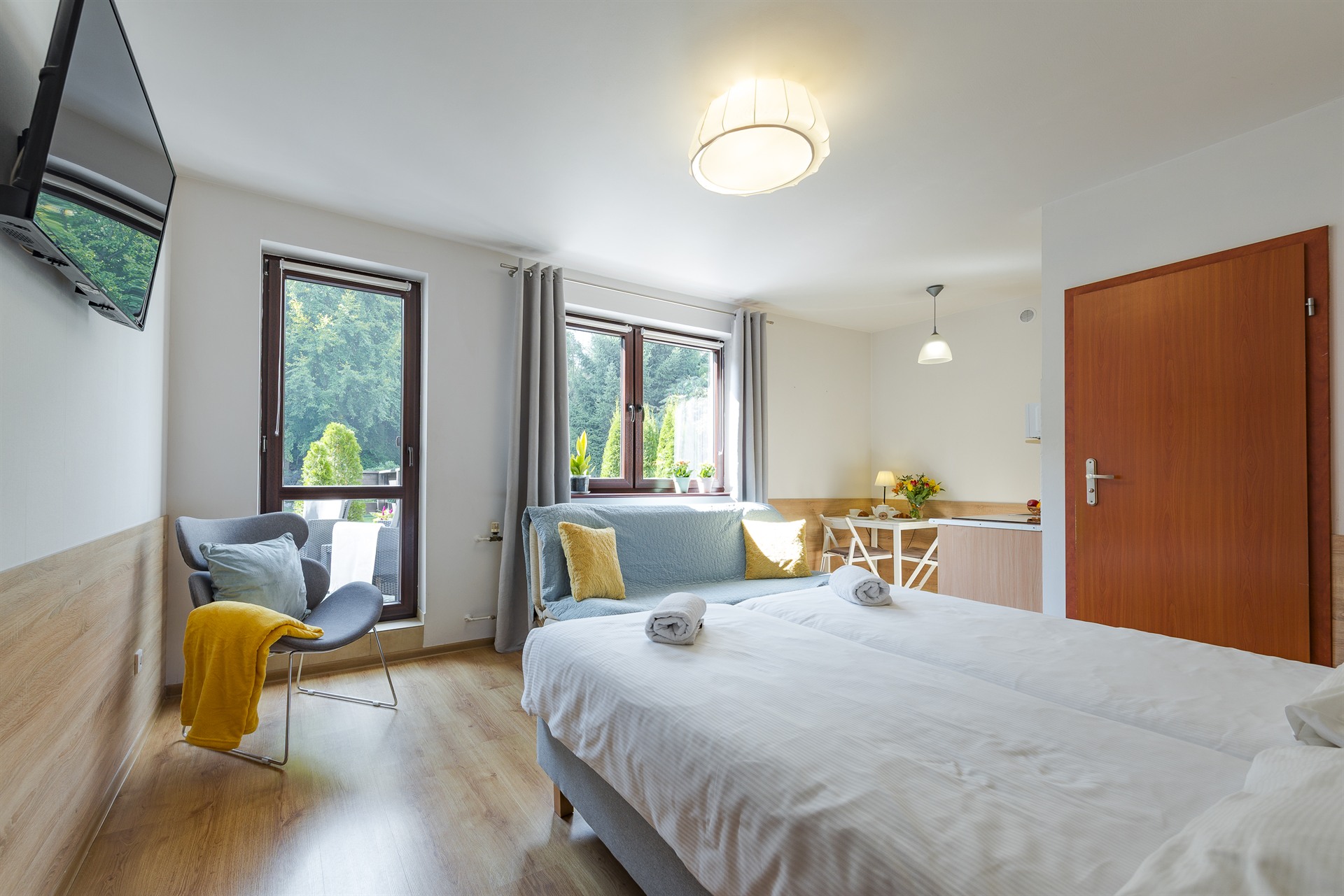 Jasny pokój hotelowy z dwoma łóżkami, niebieską sofą, żółtymi poduszkami i fotelem z żółtym kocem