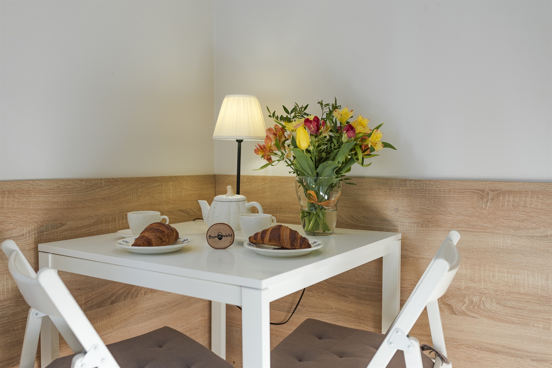 Biały stolik z czajnikiem, dwiema filiżankami, dwoma croissantami i bukietem żółto-różowych kwiatów